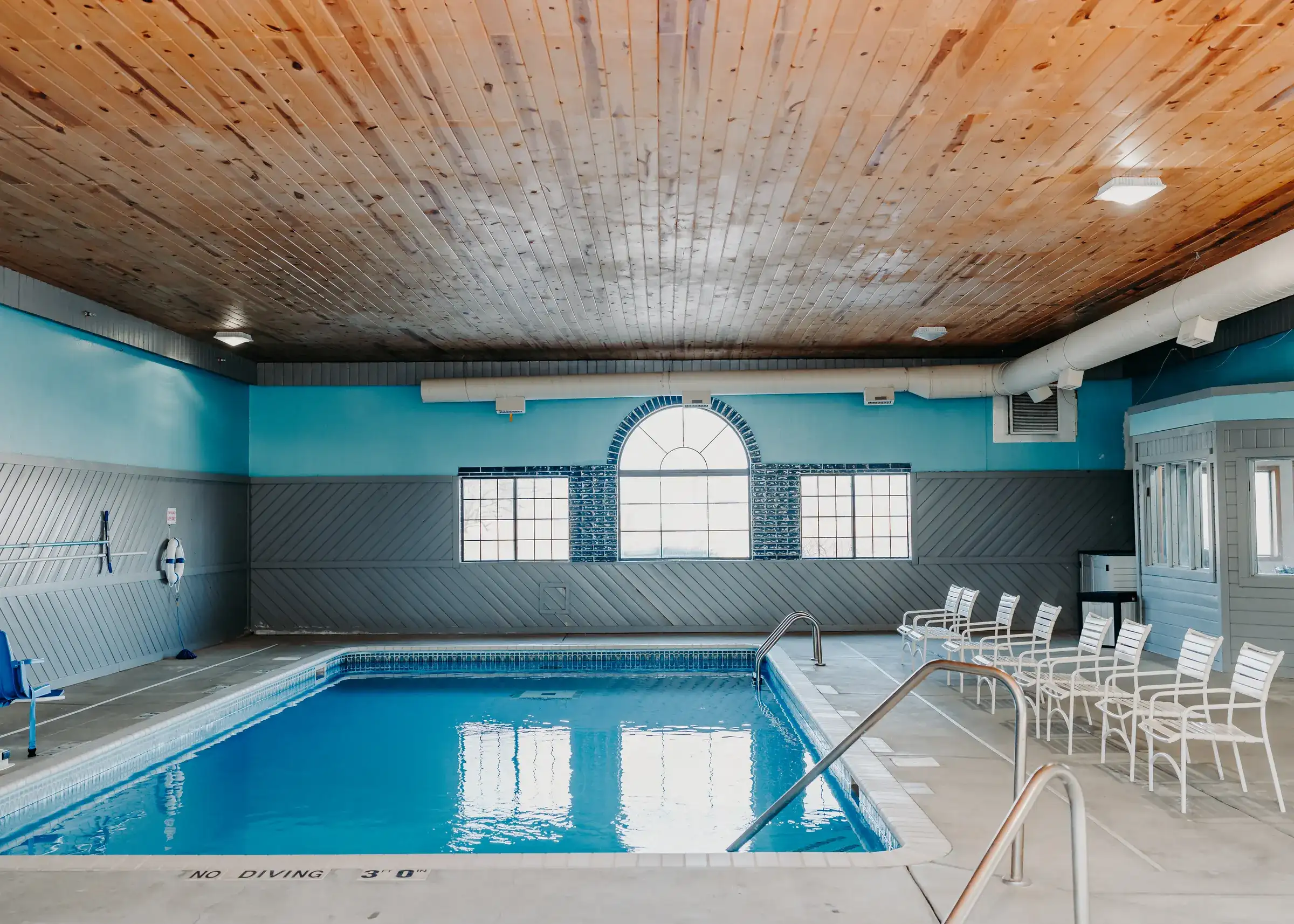 The pool room's arched window wall and pine plank ceiling, with loungers along the far side